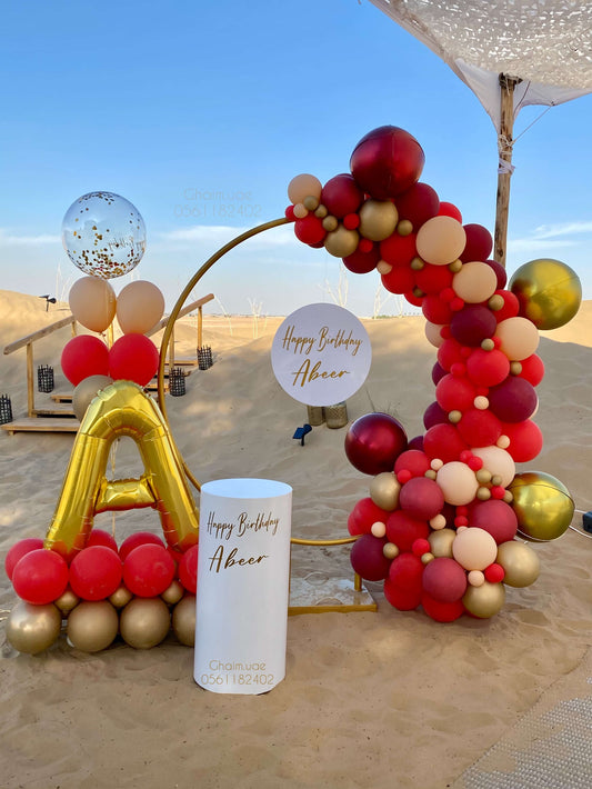 Red and gold birthday balloon backdrop with circular frame, letter balloon, and custom text sign in desert setting