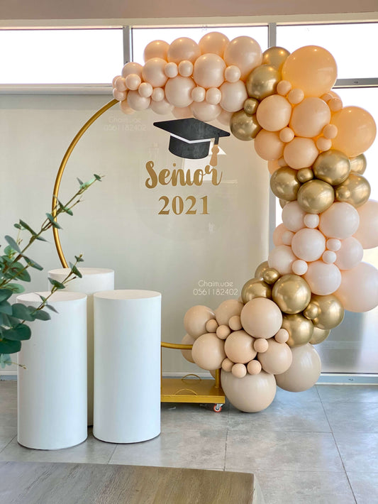 "Elegant graduation balloon arch setup with nude, white, and gold balloons on a golden round backdrop, featuring a transparent board with 'Senior 2021' and a graduation cap, next to three white display stands."