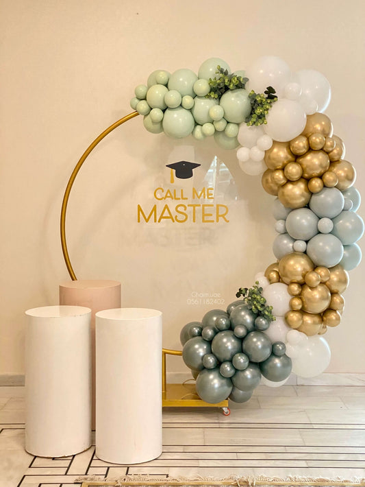 "Graduation balloon arch in soft green, white, and gold colors on a golden round backdrop with greenery accents. A clear board displays the phrase 'Call Me Master' with a graduation cap, next to white display stands."