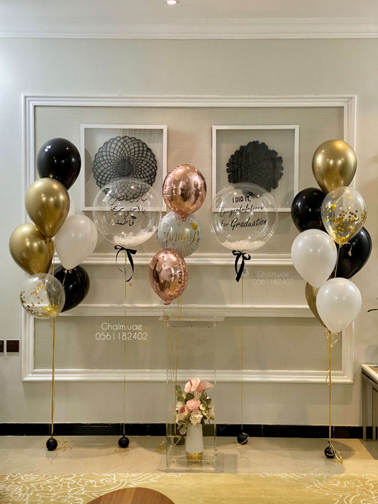 "Elegant graduation helium balloon bouquet featuring black, gold, white, rose gold, and transparent balloons with congratulatory messages in English and Arabic, arranged symmetrically around a clear podium with a floral arrangement."