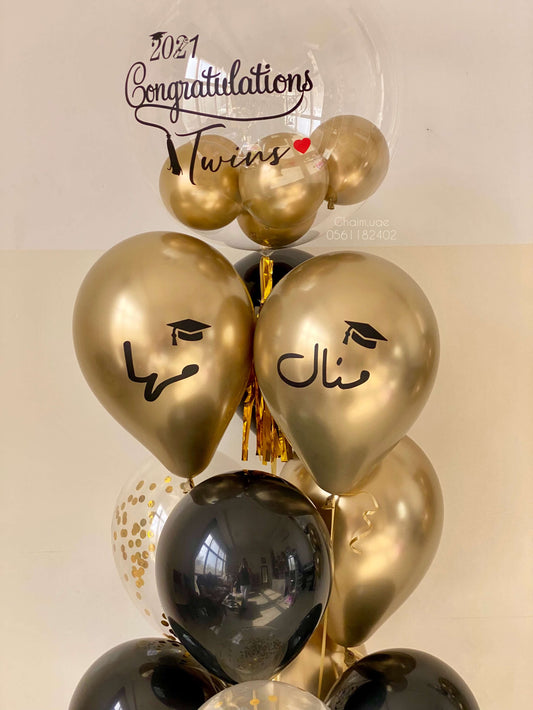 "A graduation balloon arrangement for twins featuring gold, black, and transparent balloons. The top clear balloon says 'Congratulations Twins 2021' with a red heart and graduation cap, while two gold heart-shaped balloons display the Arabic names 'Shatha' and 'Manal' with graduation cap icons."