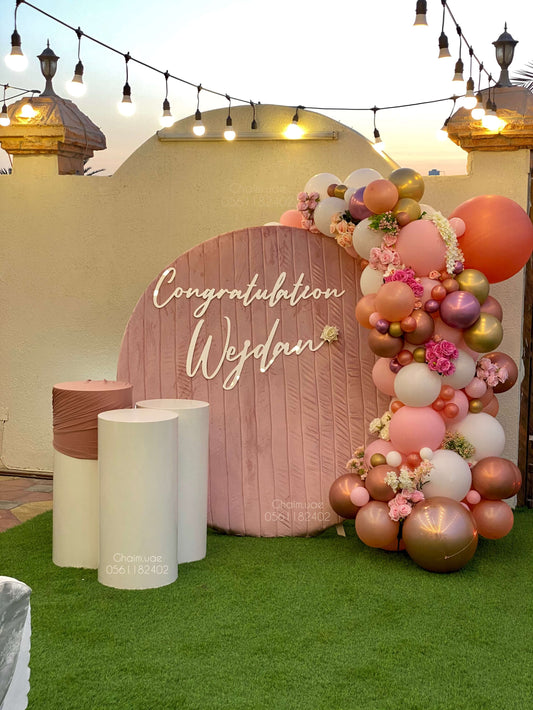 "Outdoor celebration setup with a round blush pink backdrop reading 'Congratulations Wesdan', decorated with a balloon garland in rose gold, white, pink, and floral accents, next to three white display stands on artificial grass."
