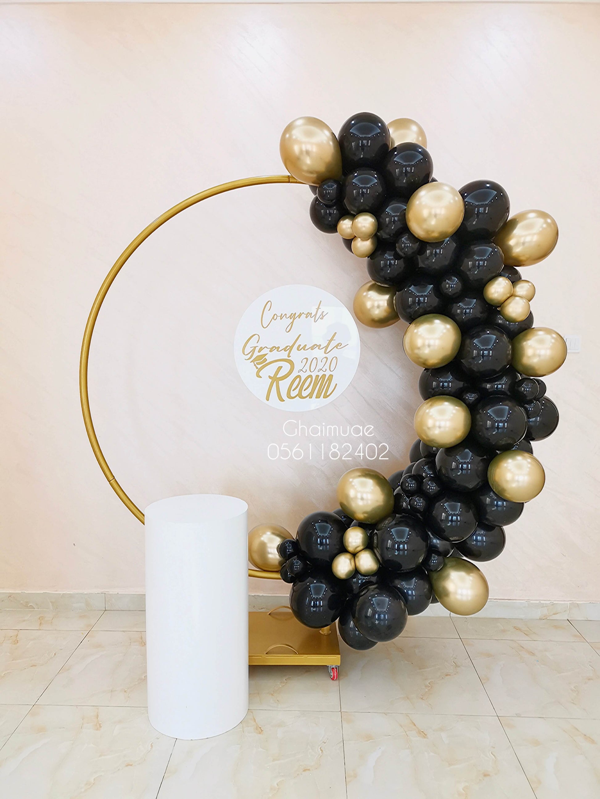 "Graduation balloon arch in black and gold colors on a golden round backdrop, featuring a personalized clear board with 'Congrats Graduate ' and a white display stand."