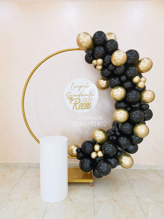 "Graduation balloon arch in black and gold colors on a golden round backdrop, featuring a personalized clear board with 'Congrats Graduate ' and a white display stand."