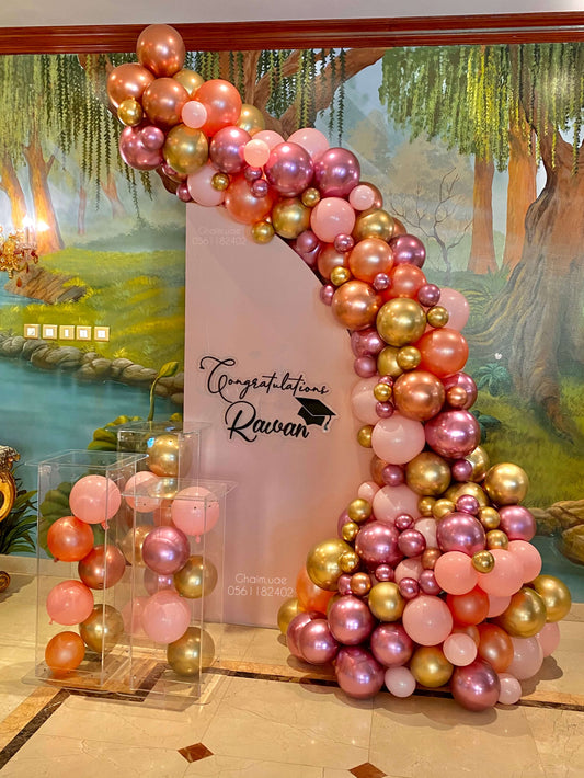 "Colorful graduation balloon setup with a pink, rose gold, and gold balloon garland on a curved backdrop, featuring a custom blush backdrop with 'Congratulations Rawan' text and two clear boxes filled with matching balloons."