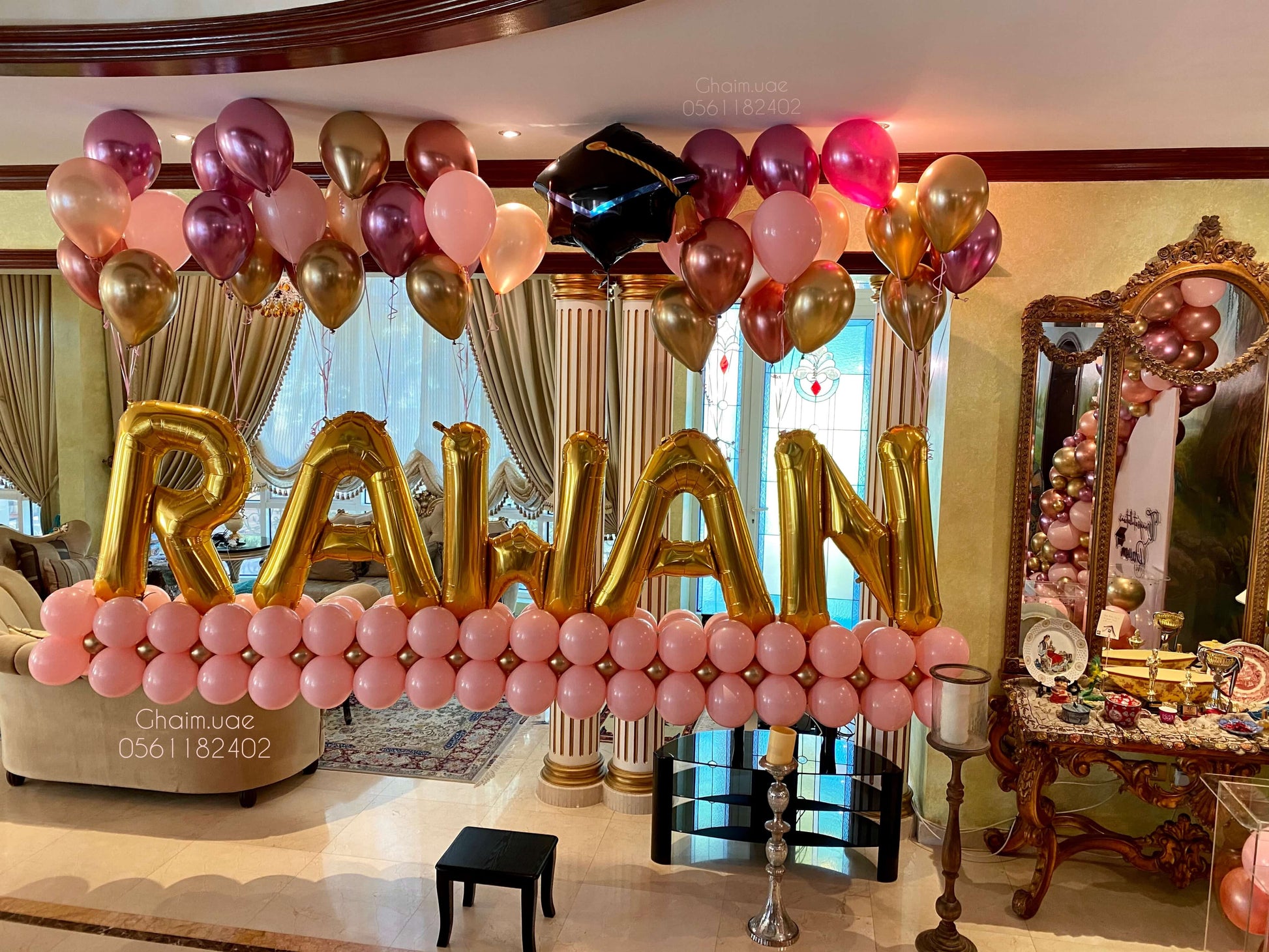 "Graduation balloon display featuring large gold letter balloons spelling 'RAWAN', arranged on a pink balloon base, with floating rose gold, pink, and gold helium balloons and a black graduation cap balloon above."