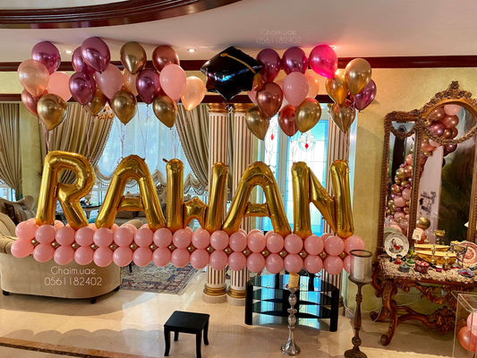 "Graduation balloon display featuring large gold letter balloons spelling 'RAWAN', arranged on a pink balloon base, with floating rose gold, pink, and gold helium balloons and a black graduation cap balloon above."