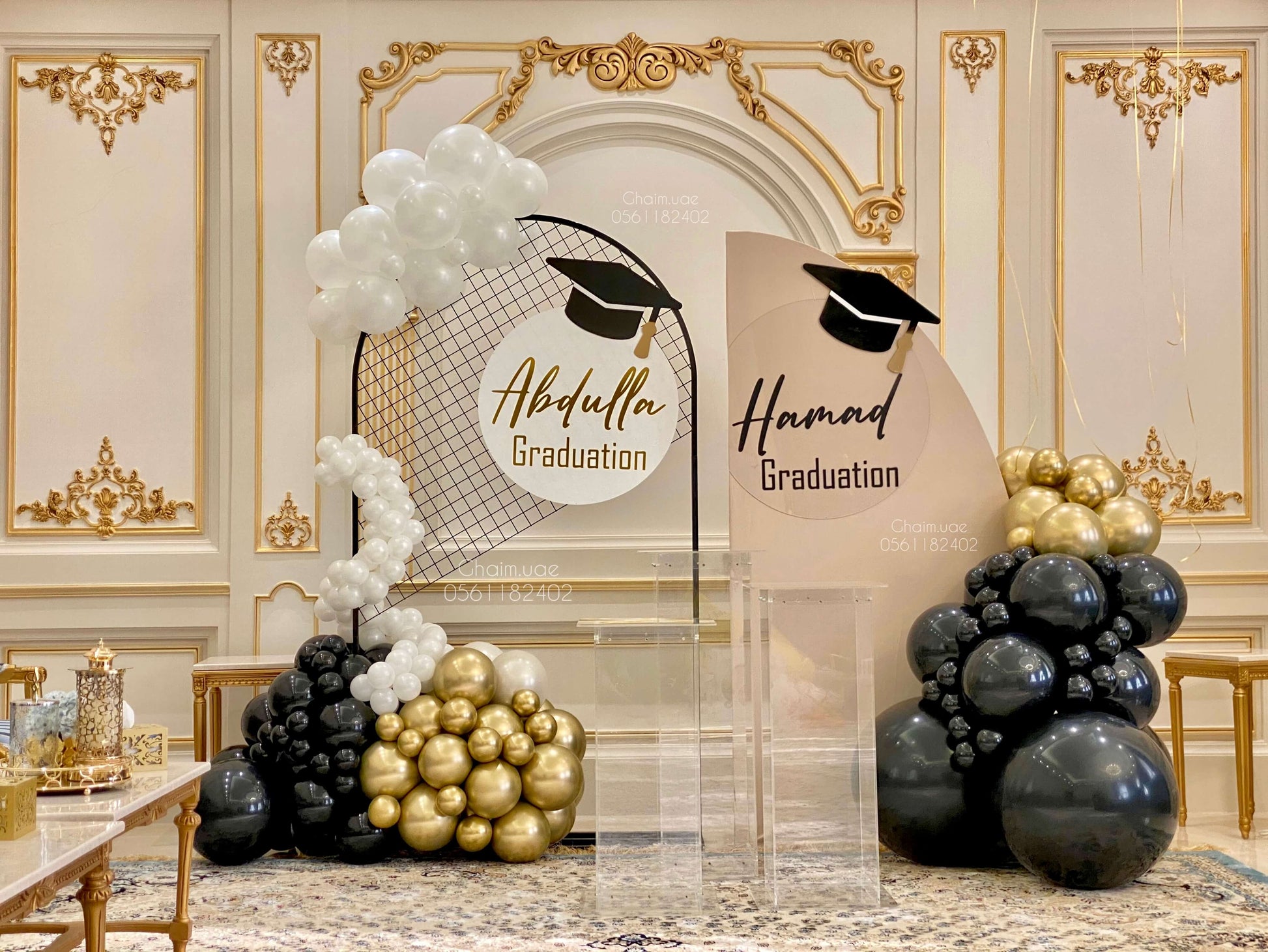 "Elegant indoor graduation setup featuring two custom backdrops for 'Abdulla' and 'Hamad' with graduation caps, decorated with black, gold, and white balloons, and clear acrylic stands in a luxurious hall."