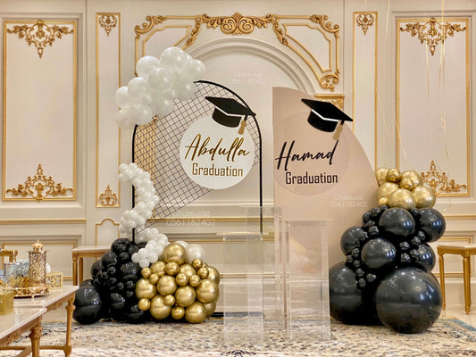 "Elegant indoor graduation setup featuring two custom backdrops for 'Abdulla' and 'Hamad' with graduation caps, decorated with black, gold, and white balloons, and clear acrylic stands in a luxurious hall."
