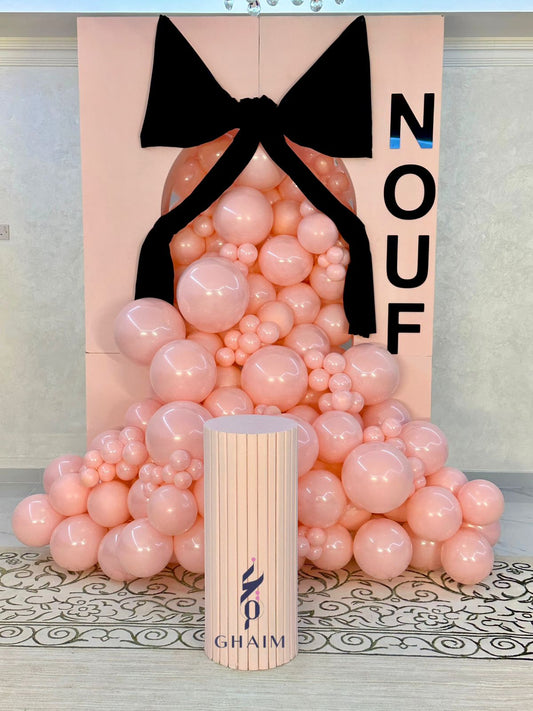 Signature pink balloon setup with a pastel backdrop, oversized black bow, personalized name “NOUF”, and a ribbed pedestal
