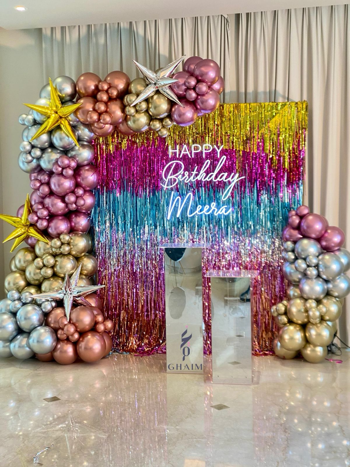 Glamorous birthday backdrop with metallic balloon garland, colorful fringe curtain, star-shaped balloons, neon sign, and clear acrylic stands