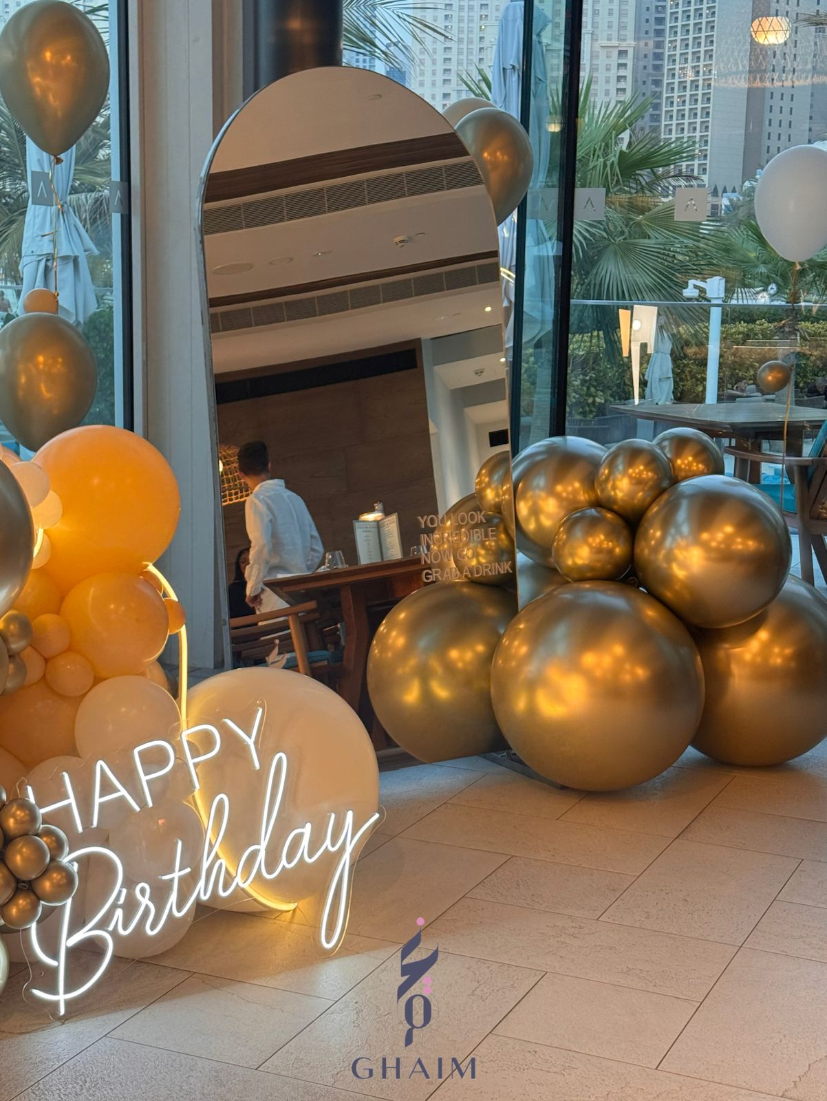Luxurious golden birthday setup with large gold balloons, a neon “Happy Birthday” sign, arched mirror, and floating helium balloons