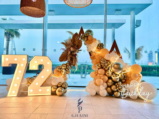 Luxury birthday decor with gold and white balloon garland, triangular wooden backdrops, pampas floral accents, LED string lights, “Happy Birthday” neon sign, and marquee numbers 72