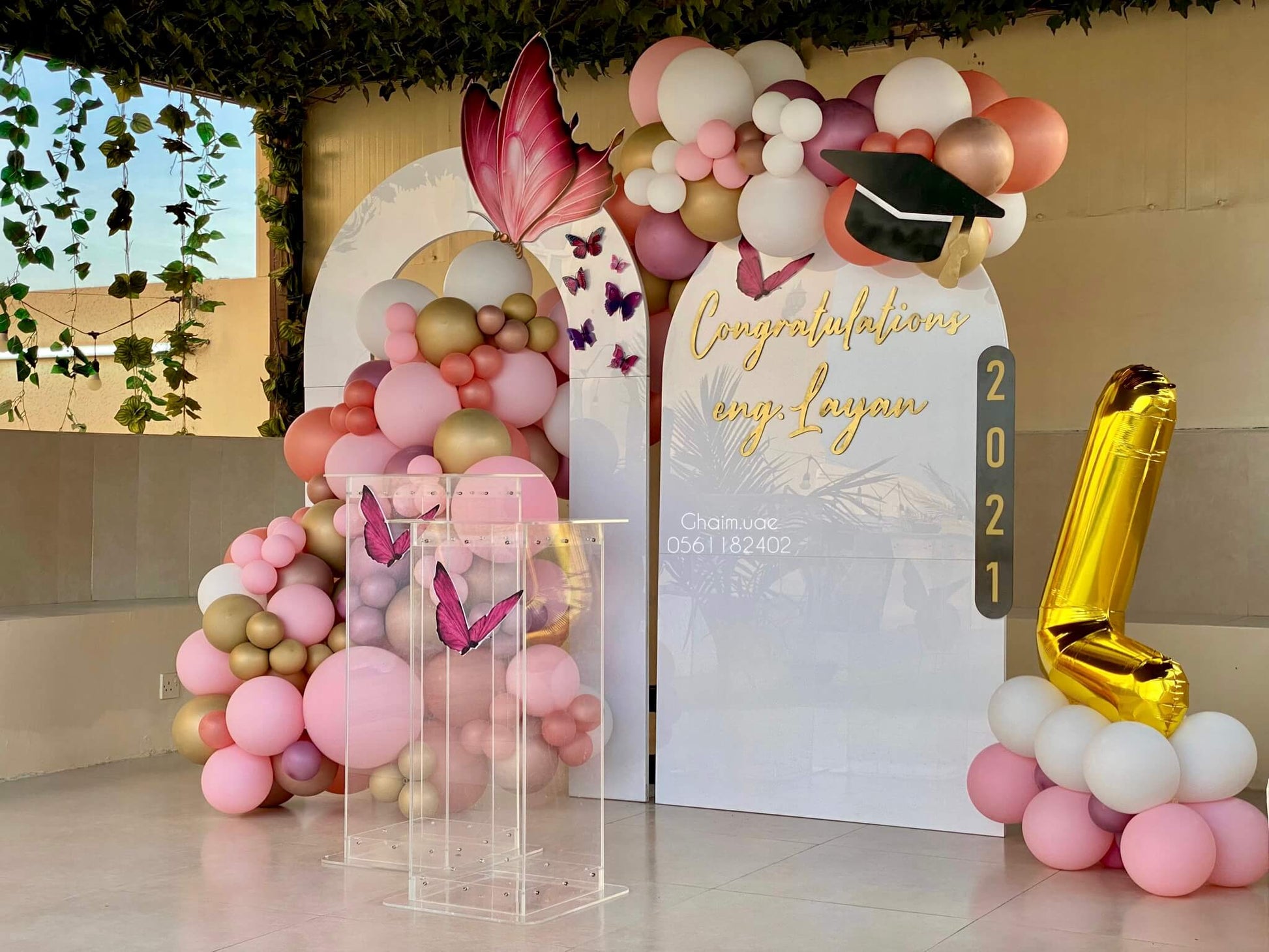 "Outdoor graduation balloon setup for Eng. Layan featuring pink, white, and gold balloon garlands, butterfly decorations, a custom backdrop with 'Congratulations Eng. Layan' and a large gold number 1 balloon."