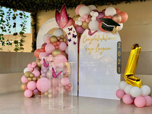 "Outdoor graduation balloon setup for Eng. Layan featuring pink, white, and gold balloon garlands, butterfly decorations, a custom backdrop with 'Congratulations Eng. Layan' and a large gold number 1 balloon."